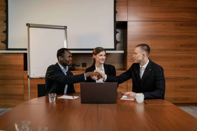 Two men shake hands in a boardroom while a woman looks on